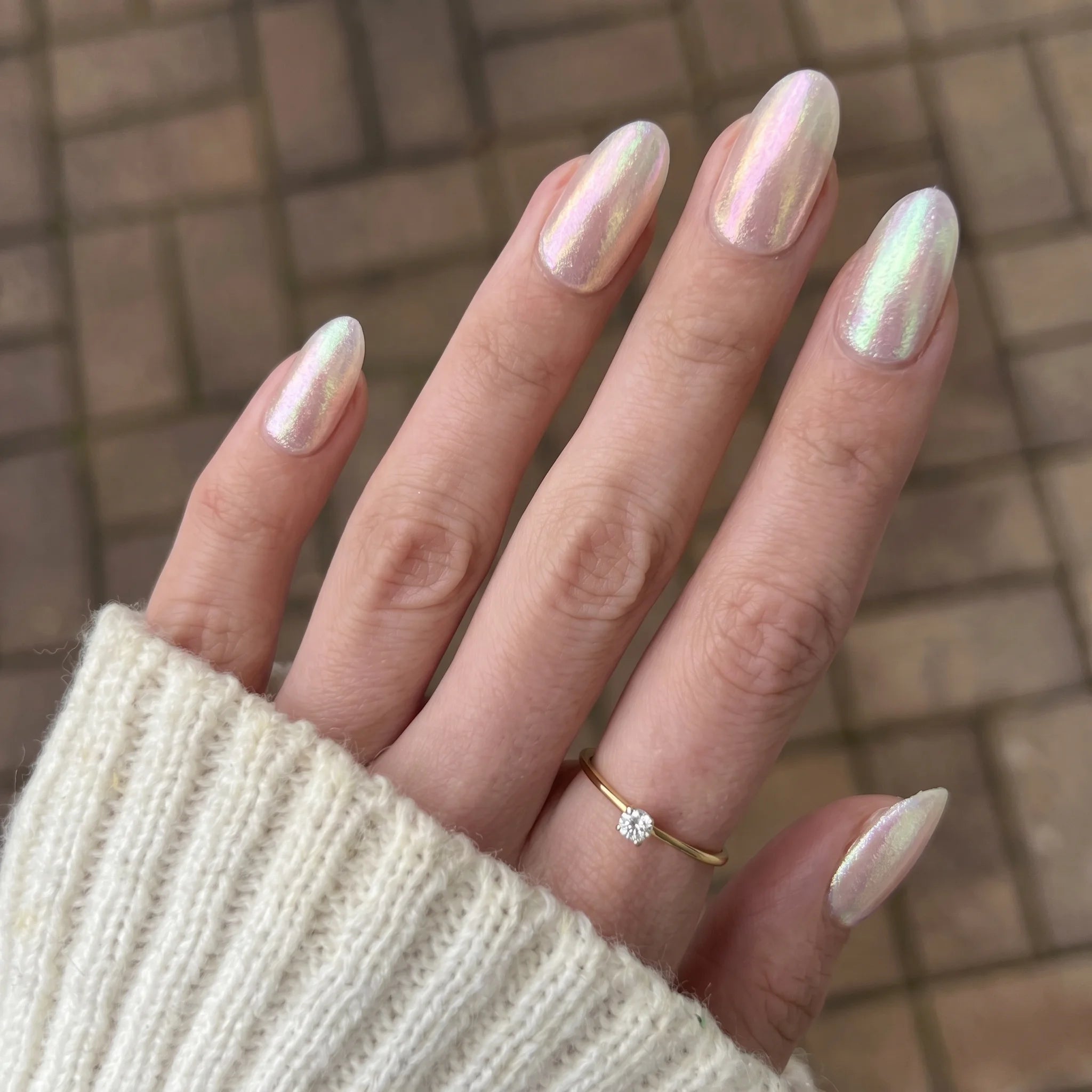 Een hand met lange amandelvormige nagels in glinsterende iriserende witte Cosmic Shift chrome poeder en een gouden ring
