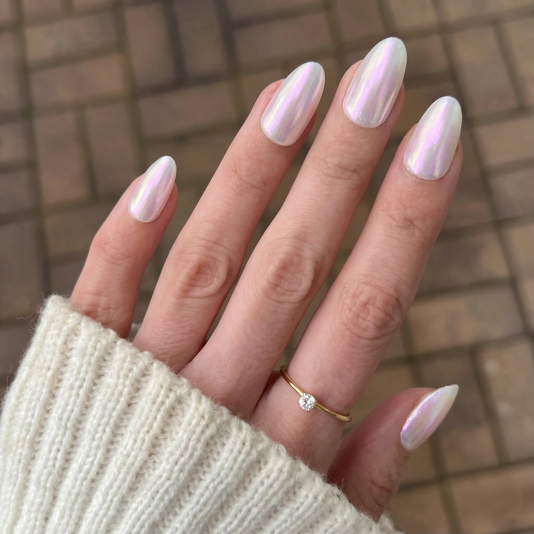 Hand met lange amandelvormige nagels in glanzend parelmoerwit crimson shift chrome powder en gouden ring met diamant
