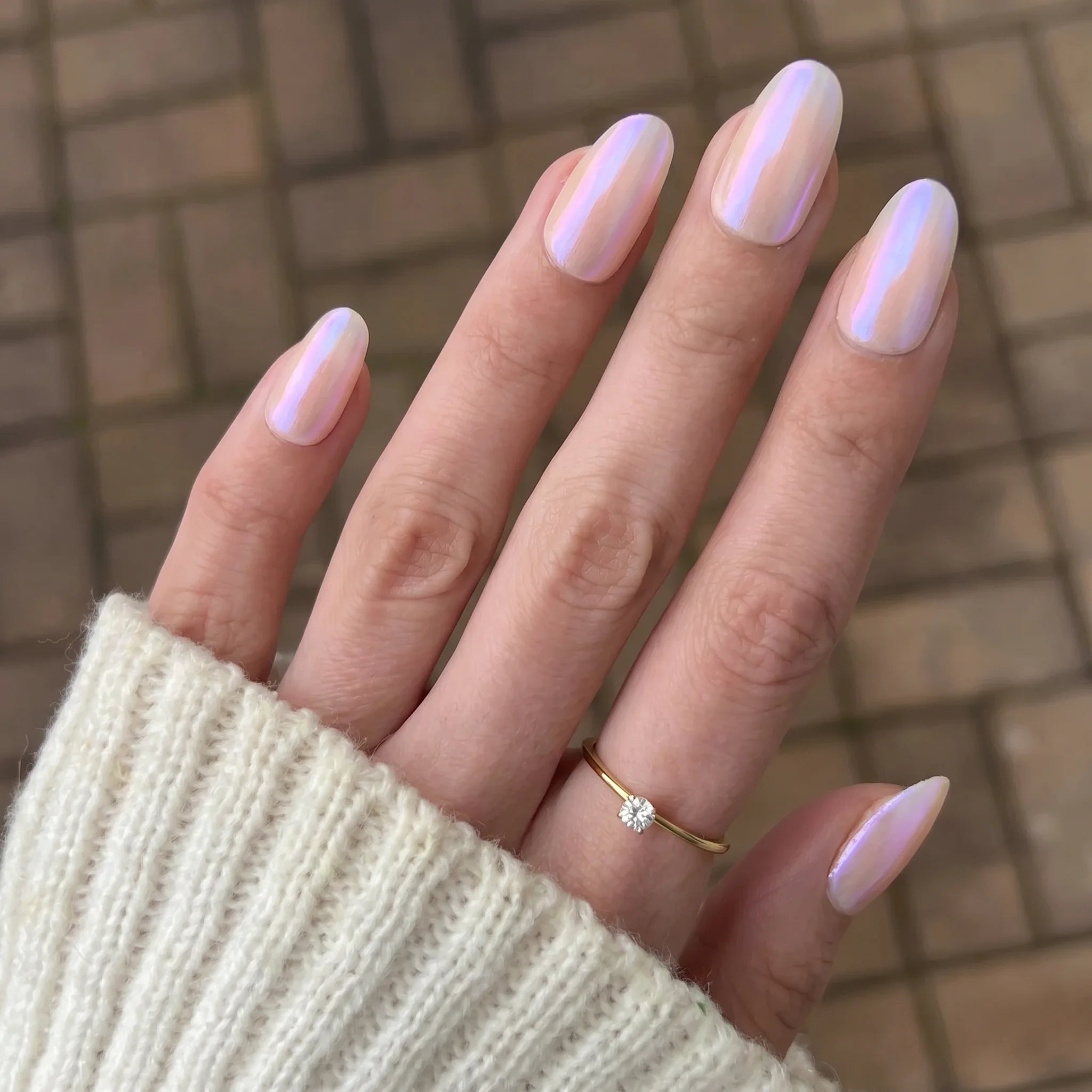Een hand met lange amandelvormige nagels in zacht parelmoer roze holografische emerald shift chrome poeder polish en fijne gouden ring