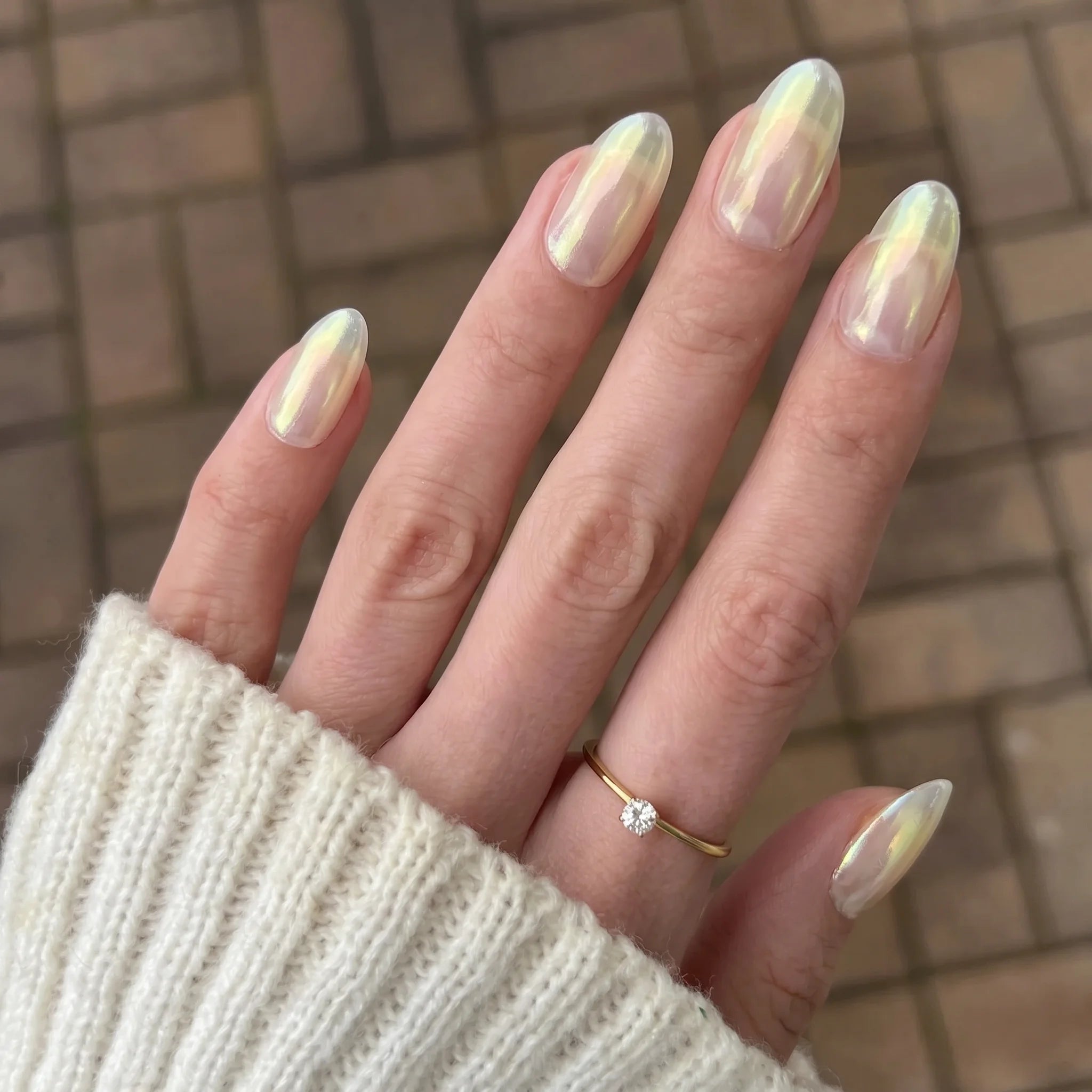 Een hand met lange amandelvormige nagels met een glinsterende holografische sapphire shift chrome poeder finish en een fijne gouden ring met een heldere steen