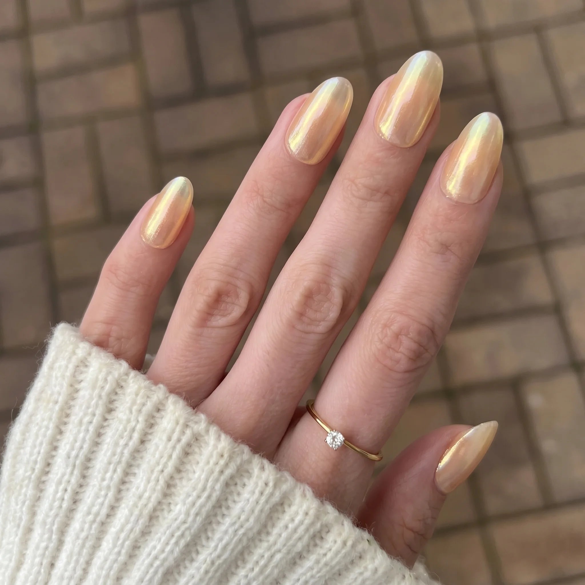 Een hand met lange amandelvormige nagels gelakt in glanzende goud-iriserende sapphire shift chrome powder polish met een delicate diamant ring